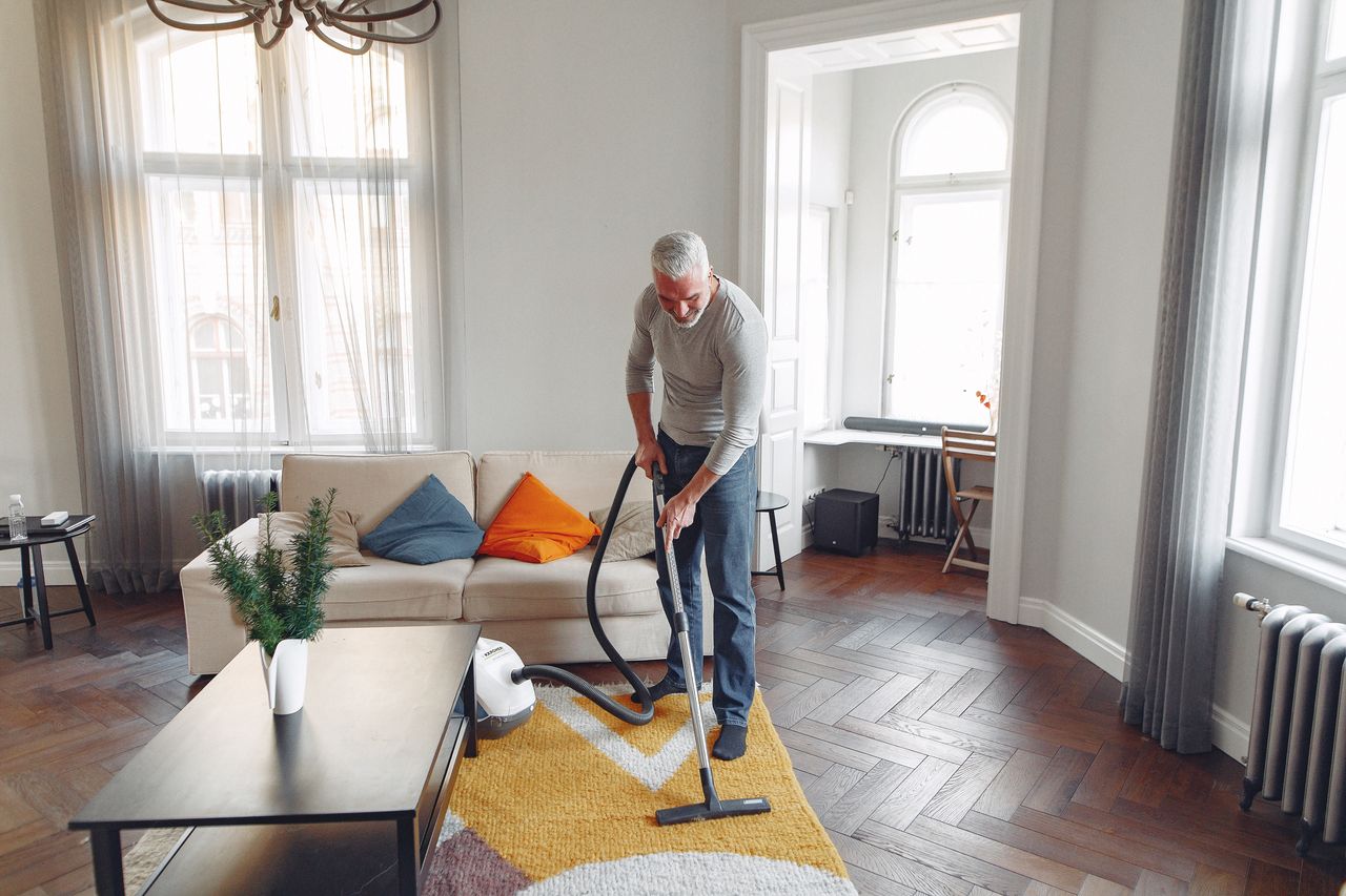 services-01 From above of focused grey haired man with beard vacuum cleaning carpet at home